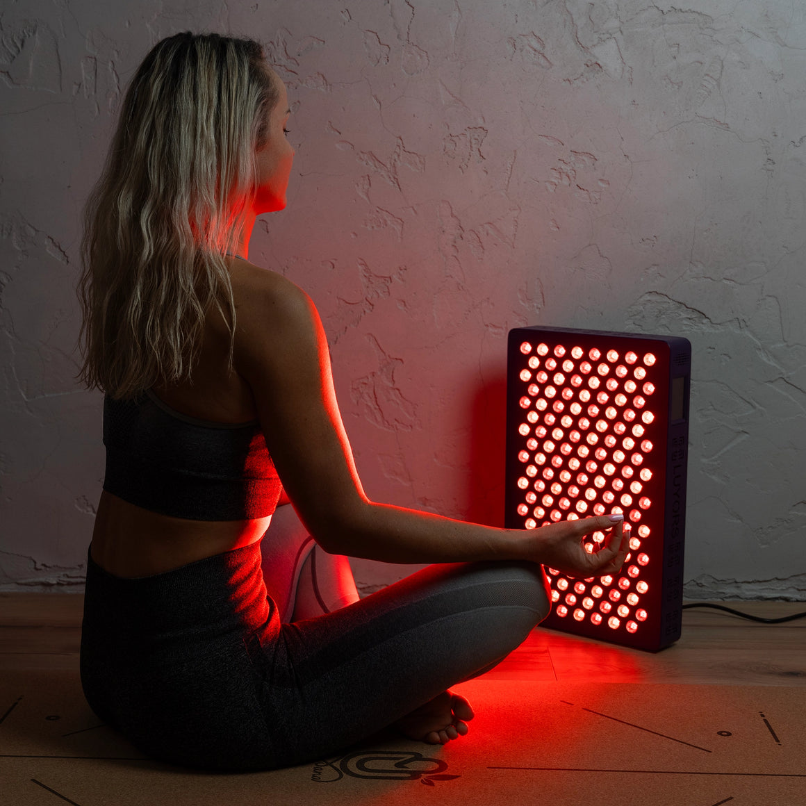 Woman-sitting-cross-legged-on-a-yoga-mat-with-her-back-lit-by-the-6-Wavelength-LED-Therapy-Panel's-red-light