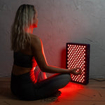 Woman-sitting-cross-legged-on-a-yoga-mat-with-her-back-lit-by-the-6-Wavelength-LED-Therapy-Panel's-red-light